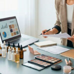 A professional beauty blogger and business owner working at a desk with makeup palettes, skincare bottles, a laptop showing analytics, and a camera setup for social media content creation.