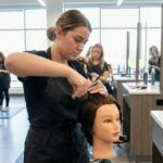 A cosmetology student practicing hair cutting techniques on a mannequin head in a modern beauty school classroom with other students training in the background.