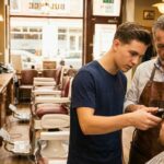 A seasoned barber mentor in a leather apron guides a student's hand holding clippers during hands-on barber training on a client in a classic barbershop.