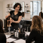 Beauty instructor demonstrating a hands-on hair technique to adult students in a professional training classroom with mannequin heads, lesson papers, mirrors, and salon tools.