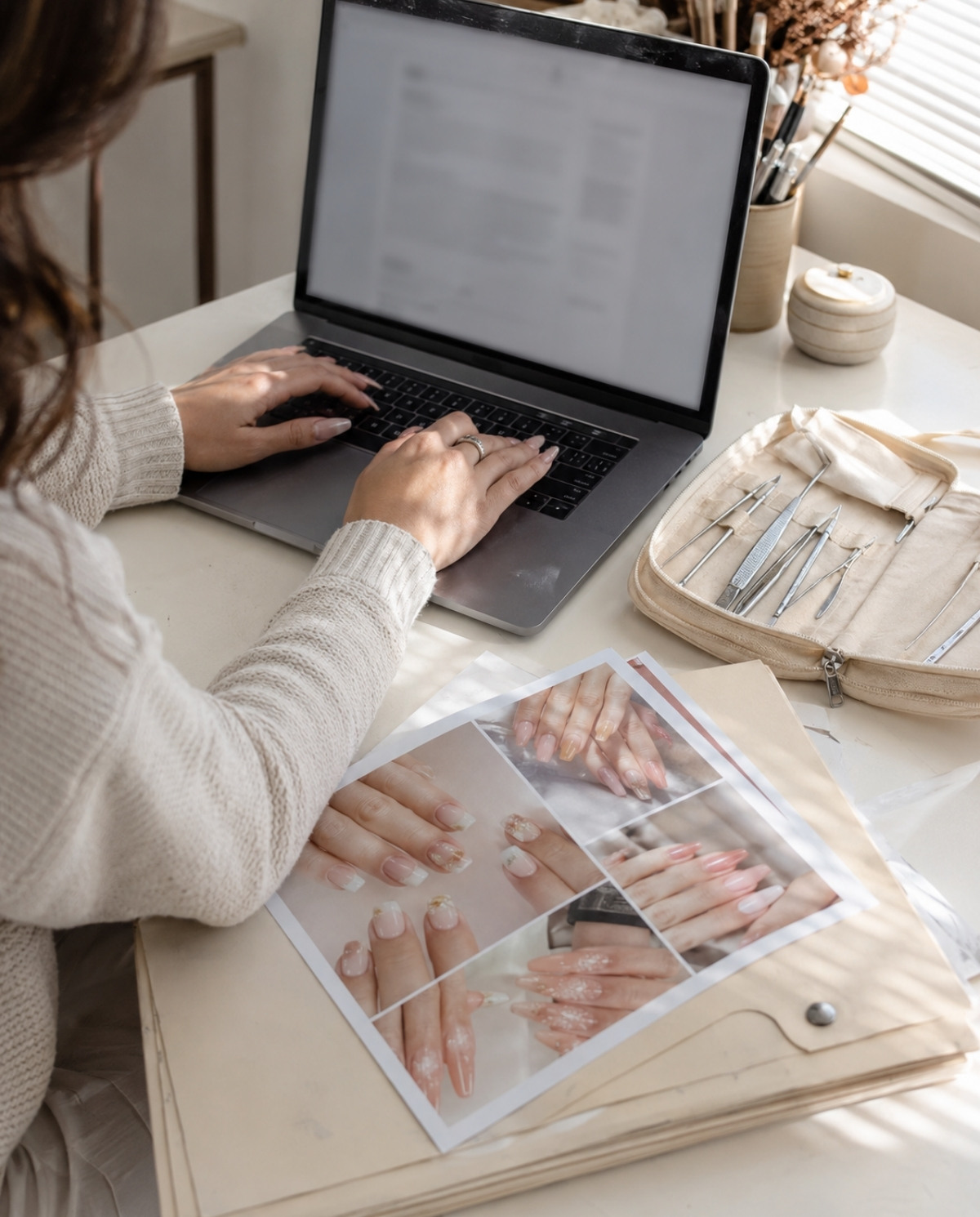 Beginner nail technician editing a resume on a laptop beside printed manicure photos, a license folder, and clean tools.