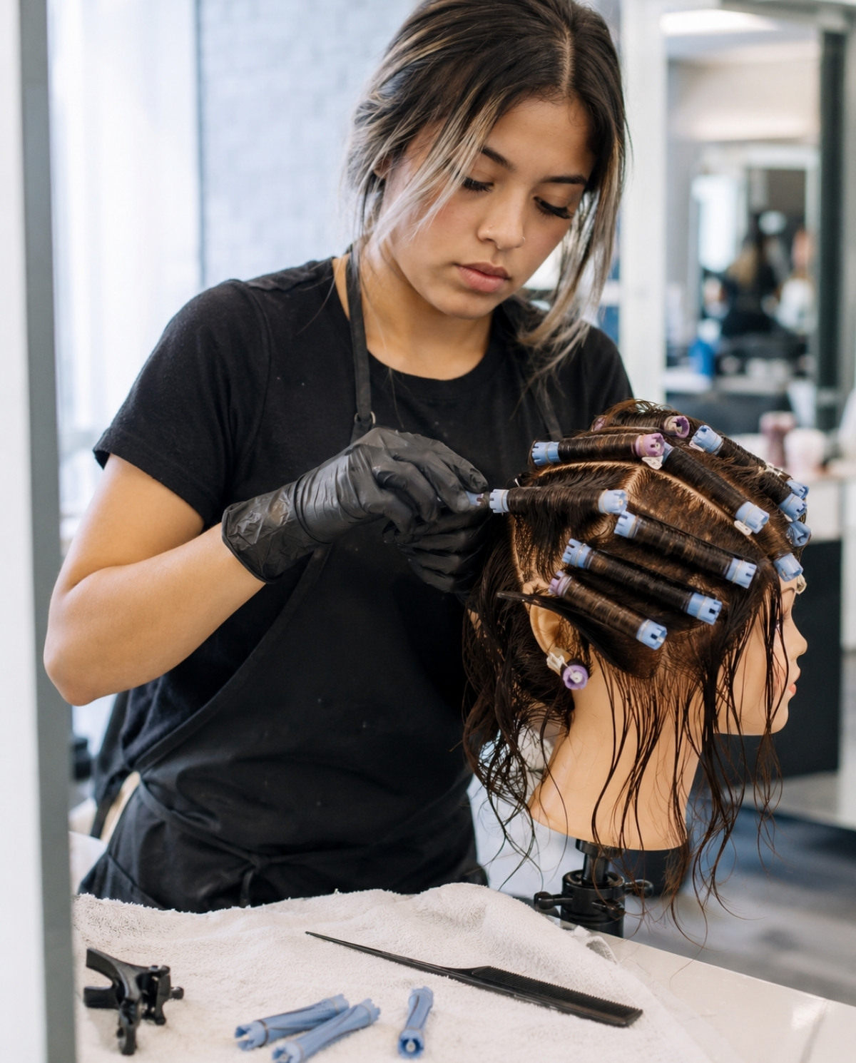 Cosmetology student wearing gloves practices chemical texture preparation by carefully wrapping sectioned damp mannequin hair around perm rods.