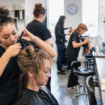 Cosmetology student styles an adult model while classmates practice haircutting, hair coloring, and sanitation throughout an active training classroom.