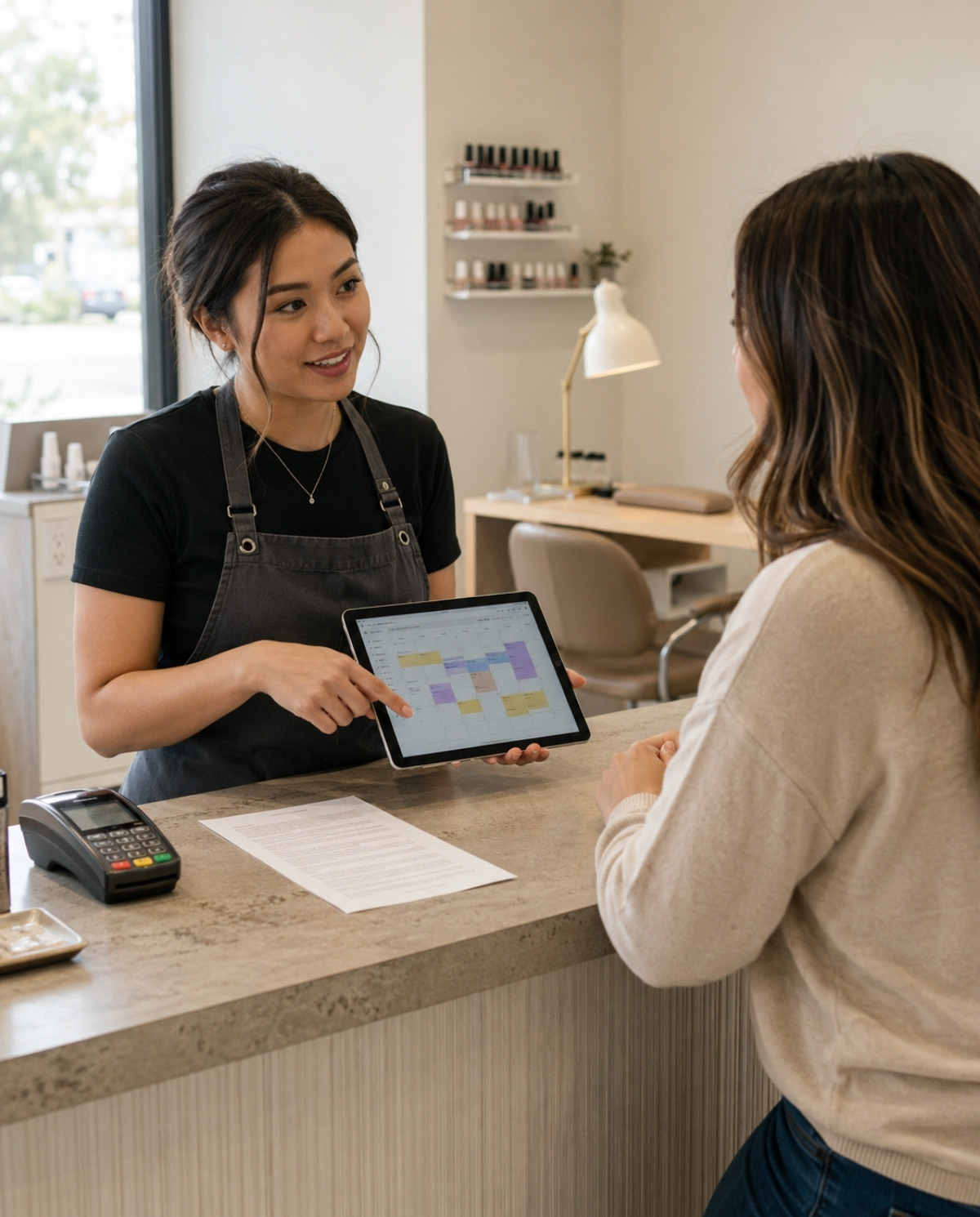 Nail professional reviews an appointment calendar with a client at reception beside a policy sheet and payment terminal with a manicure station visible in the background.