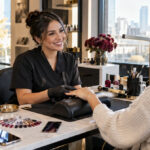Polished nail technician welcoming a returning client at a modern manicure station with nail samples, a tablet, a phone, and organized service tools.