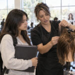 Prospective teen student and her parent observe an instructor demonstrating hair sectioning on a mannequin during a cosmetology classroom tour.