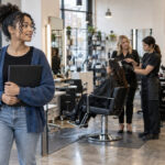 Aspiring cosmetologist holding a portfolio while observing supervised client services and structured mannequin practice in an urban training salon.